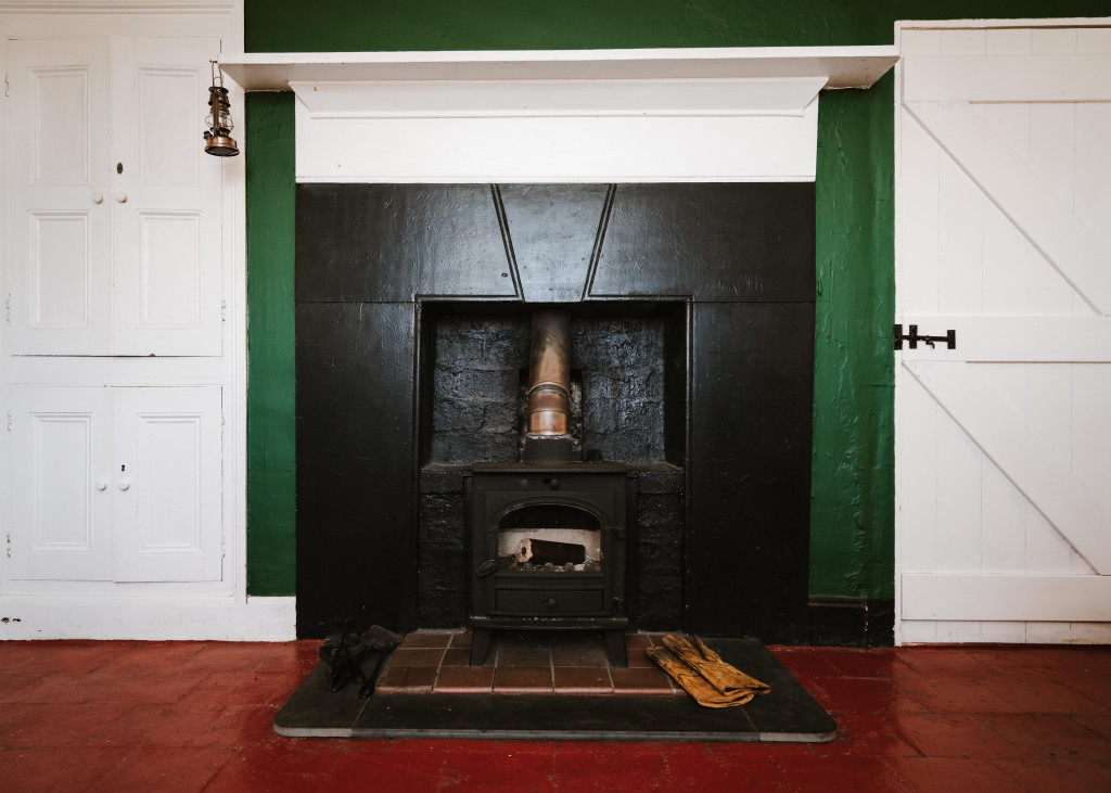 A black wood-burning stove is set in a black fireplace with a metal pipe, flanked by white cabinets and doors, against a green wall and red tiled floor.