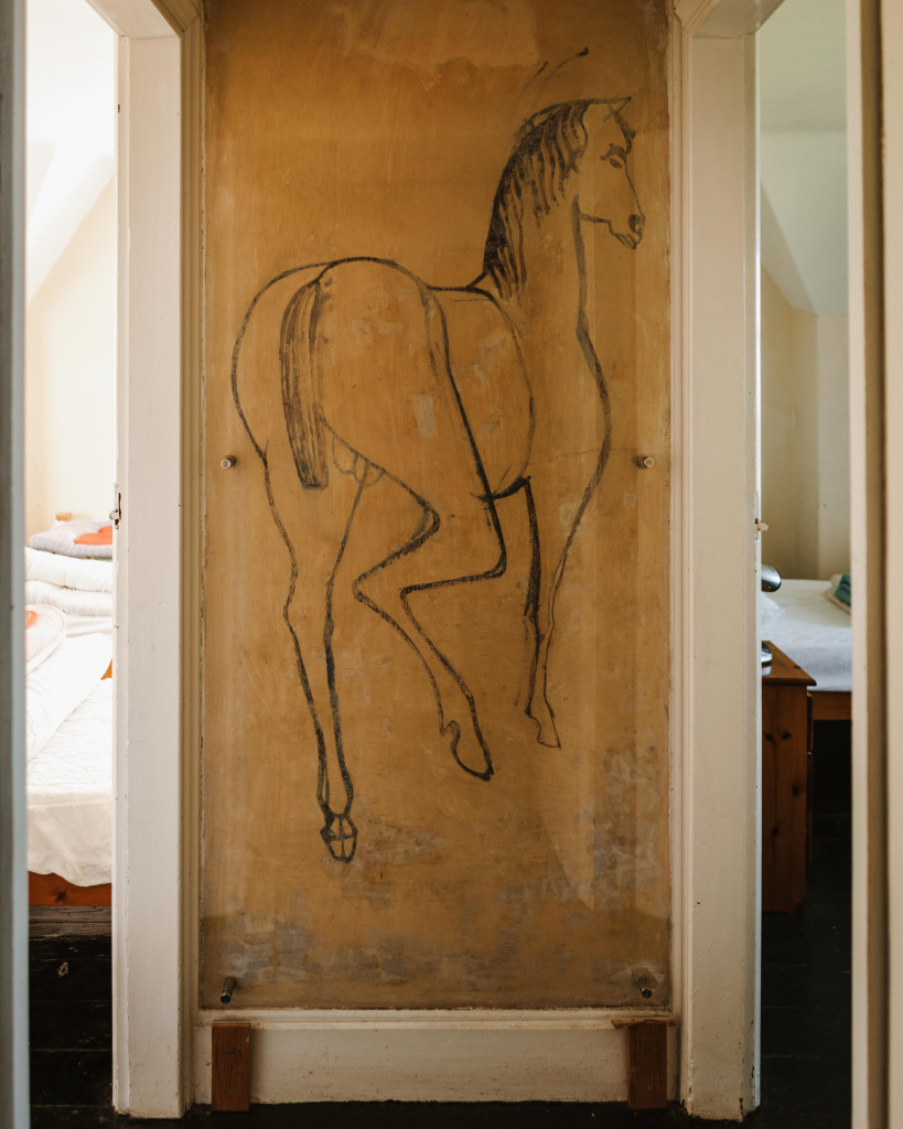 A simple horse sketch is drawn on a worn, beige wall between two doorways, with beds visible in adjoining rooms.