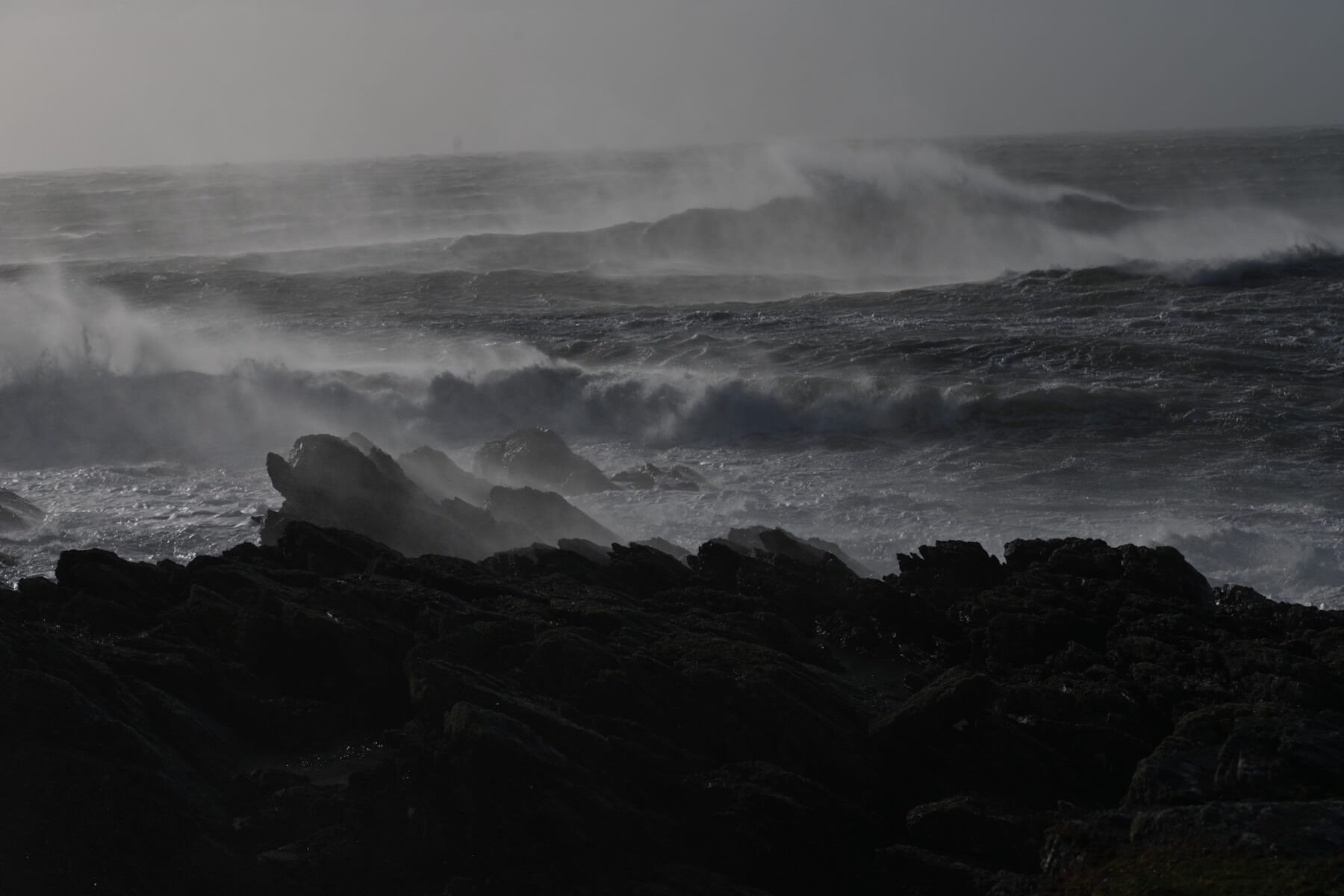 Dark, rocky shoreline in the foreground with large, crashing ocean waves and mist in the background under a cloudy sky.