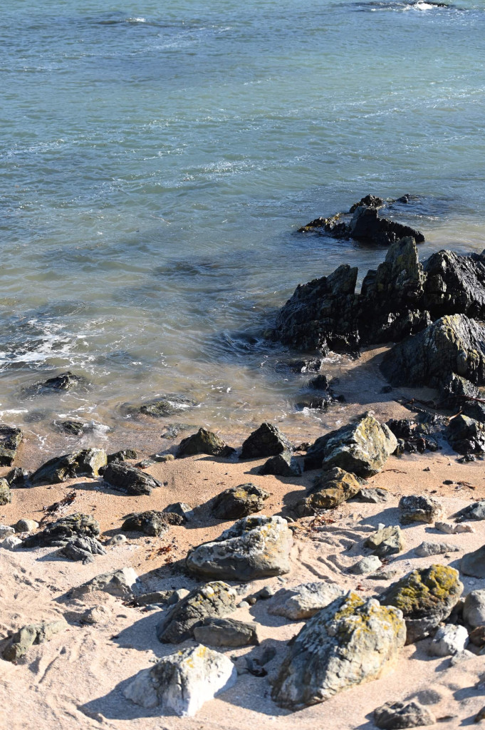 Rocks scattered on a sandy beach with small waves coming in from the blue-green ocean, echoing the serene beauty of Bardsey Island history.