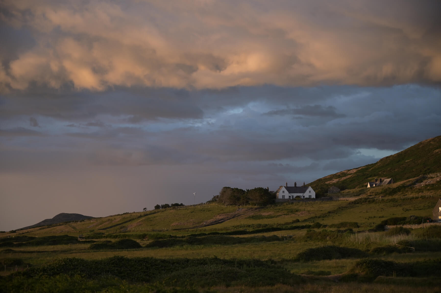 A white house sits on a grassy hillside under a dramatic cloudy sky during sunset or sunrise.