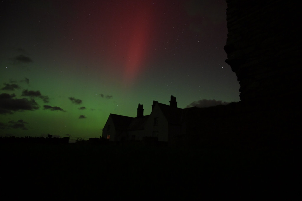 A night sky with green and red aurora lights above dark silhouettes of houses and an adjacent tall structure, evoking the tranquil landscapes often protected by Bardsey Island Trust.
