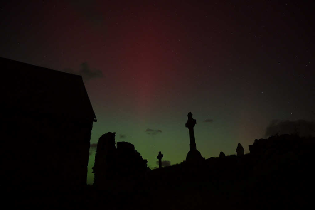 Silhouetted gravestones and a building at night on Ynys Enlli, with faint green and red aurora lights illuminating the starry sky.
