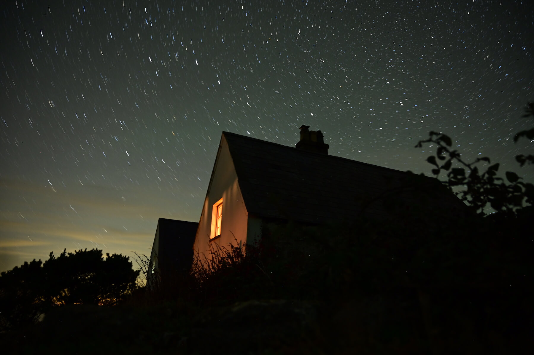 A small house with a lit window sits beneath a star-filled sky with visible star trails, nestled in shrubs and trees—a tranquil scene reminiscent of a dark sky sanctuary.