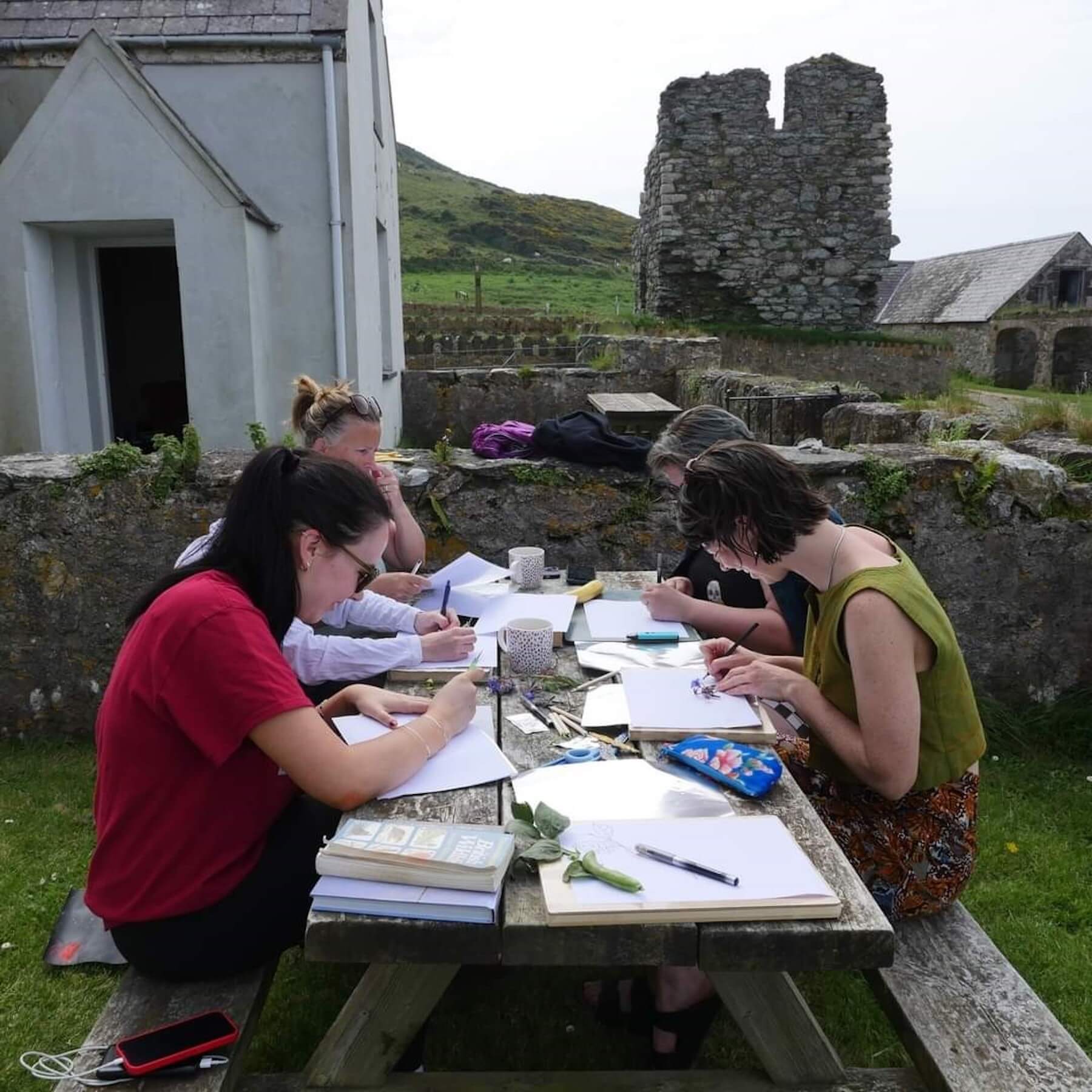 Four people sit at a wooden picnic table outdoors on Ynys Enlli, drawing or writing on paper, with ancient ruins and hills visible in the background.