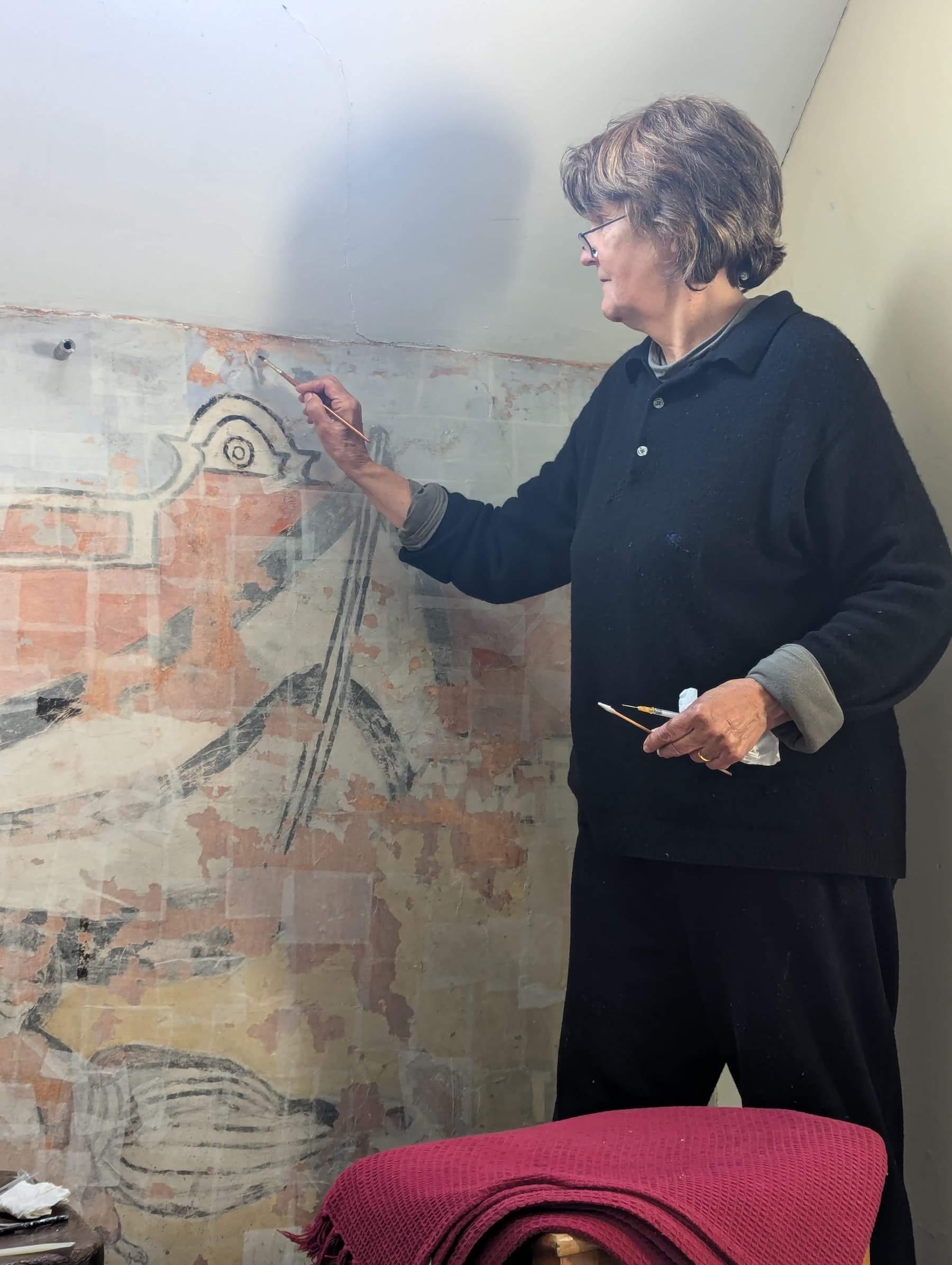 A woman stands on a step stool, carefully restoring a faded wall mural that depicts life on Enlli, with a paintbrush in one hand and restoration tools in the other.