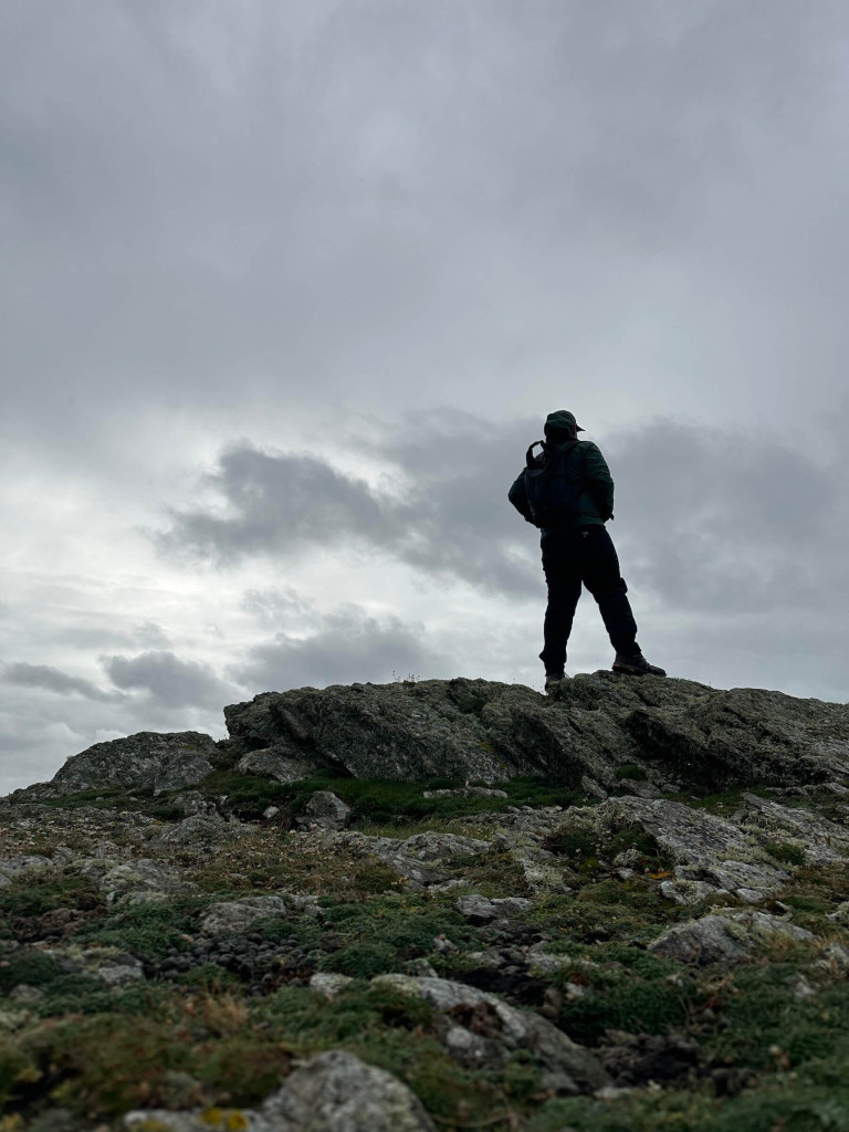 A person wearing outdoor gear stands on a rocky hilltop under a cloudy, overcast sky, gazing toward the distant outline of Ynys Enlli.