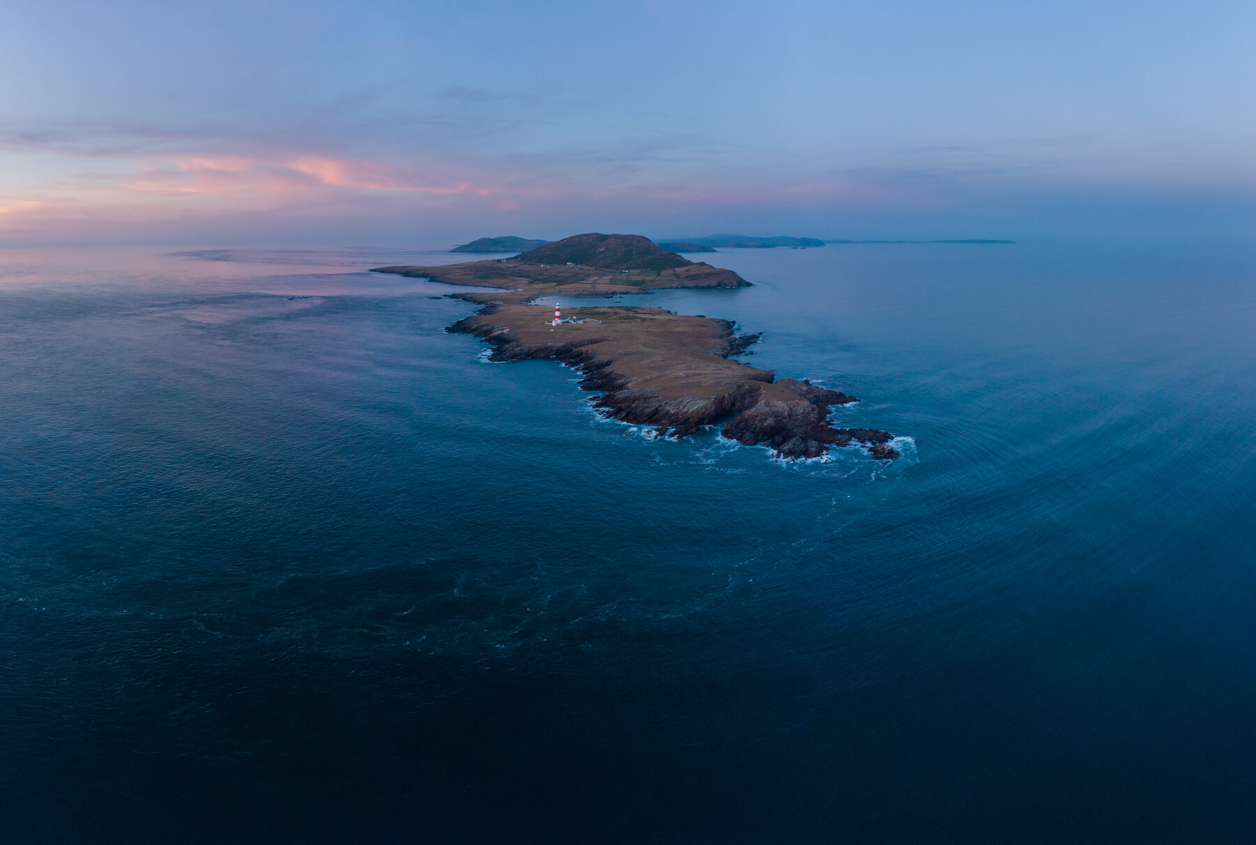 Aerial view of a narrow, rocky peninsula extending into the ocean at sunset, with low hills and scattered buildings on the land.