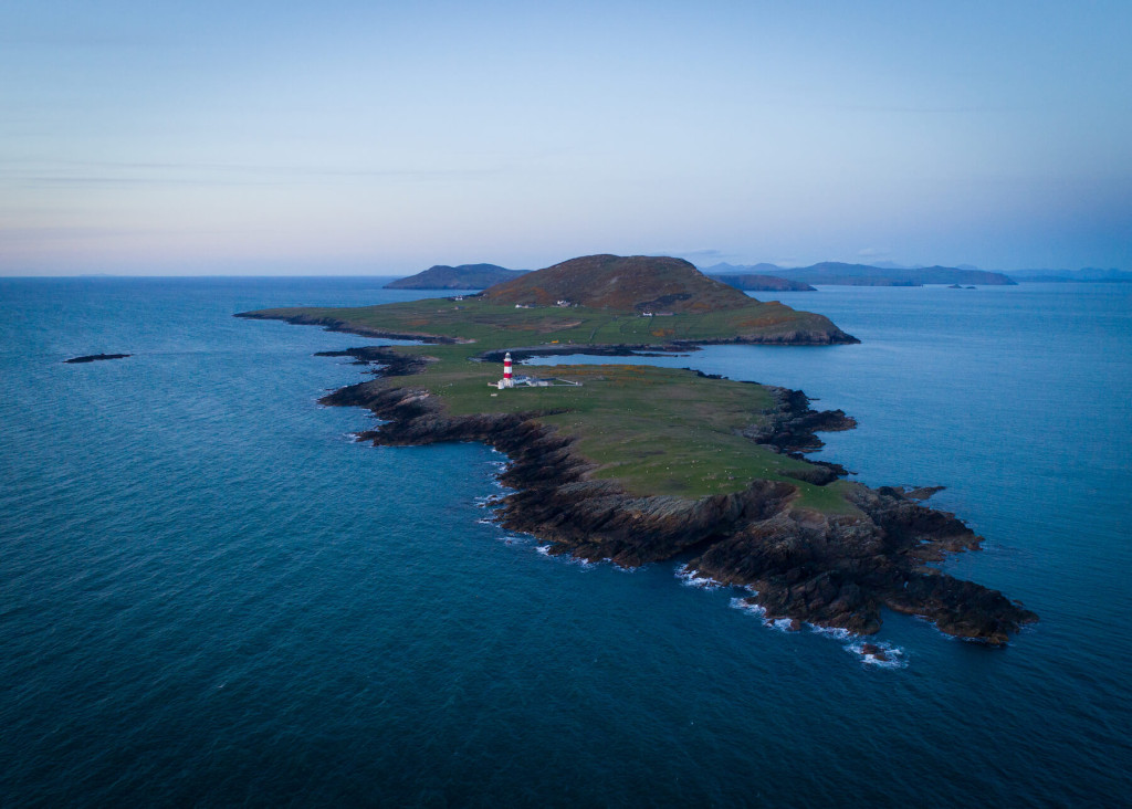 Aerial view of a narrow, rocky peninsula extending into the sea with a lighthouse near the center, surrounded by green land and distant hills—an iconic scene rich in Bardsey Island history.